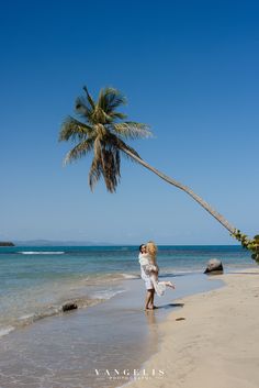 a man and woman standing on top of a sandy beach next to the ocean under a palm tree