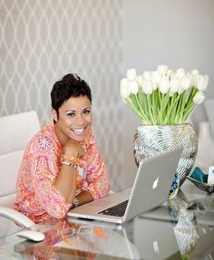 a woman sitting at a table with a laptop in front of her and flowers behind her