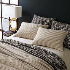 a bed with black and white comforter next to a lamp on a table in front of a window