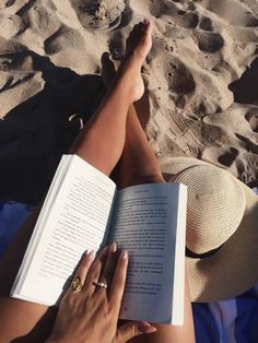 a woman laying on the beach reading a book with her feet propped up in the sand