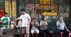 several people sitting and standing in front of a food stand on the street with signs above them