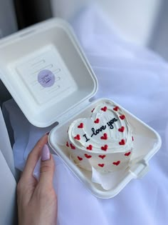 a heart shaped cake sitting in a white box on top of a bed next to a person's hand