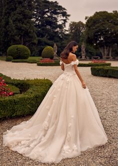 a woman in a white wedding dress standing on a gravel path with hedges and bushes behind her