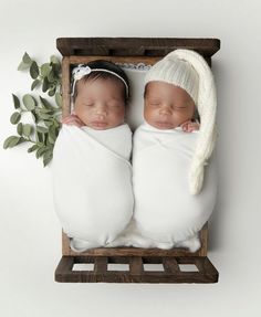 two newborn babies are sleeping in blankets and knitted hats on a wooden crate with eucalyptus leaves