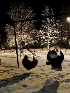 two people are playing in the snow at night with their feet up on a swing