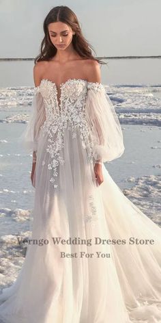 a woman standing on the beach wearing a white dress with sheer sleeves and floral appliques