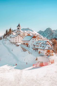 people are skiing on the snow covered slopes in front of some buildings and trees with mountains in the background