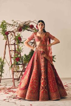 a woman in a red and gold wedding dress standing next to a floral arch with roses on it