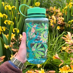 a person holding up a water bottle in front of some yellow and green flowered plants