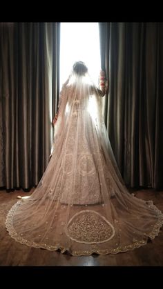 a woman in a wedding dress standing by a window with her veil pulled back and looking out the window