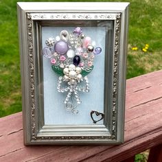 a picture frame with beads and flowers in it sitting on a wooden bench next to grass