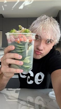 a man holding up a plastic cup filled with fruit and veggies while sitting at a table