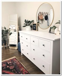 a white dresser sitting next to a mirror on top of a hard wood floor covered in plants