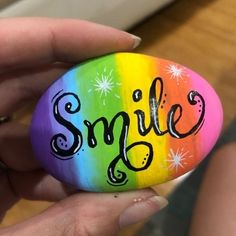 a person holding a painted rock with the word smile on it in black ink and rainbow colors