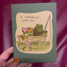 a hand holding up a card with two frogs in a boat on the water, and an i toadally love you