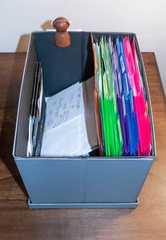 a file box filled with lots of different colored folders and pens on top of a wooden table