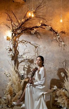 a woman sitting on a chair in front of a wall with flowers and branches hanging from it