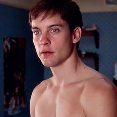 a shirtless young man standing in front of a blue wall and looking at the camera