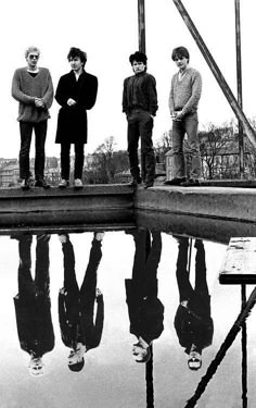 four young men standing on the edge of a body of water with their reflection in the water