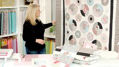 a woman pointing at a quilt on a table in front of a bookshelf