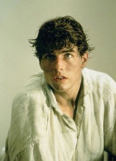 a young man with curly hair sitting in front of a white wall and looking at the camera