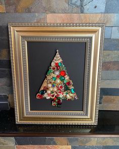 a framed christmas tree with ornaments on it in a gold and black frame against a stone wall