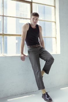 a young man leaning against a wall in front of a window with his legs crossed