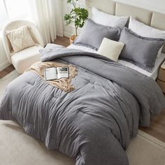 a bed with grey comforter and pillows in a room next to a chair, table and potted plant