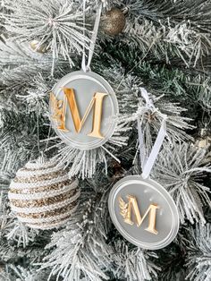 two christmas ornaments hanging from the top of a pine tree with gold letters on them