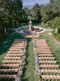 an outdoor ceremony setup with rows of wooden chairs and flowers on the grass in front of a fountain