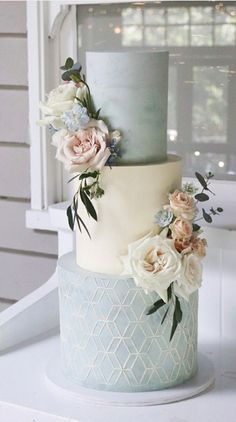 a white and blue wedding cake with flowers on the top is sitting on a table in front of a window