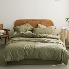 a bed with green sheets and pillows in a room next to a chair, table and potted plant