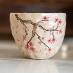 a white bowl with pink flowers painted on the side and branches in the middle, sitting on a table