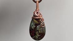 a wire wrapped pendant is hanging from a chain on a gray background with a green and red stone in the center