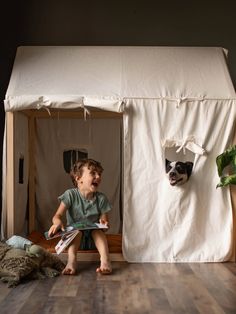 a little boy sitting on the floor in front of a tent with a dog sticking its head out