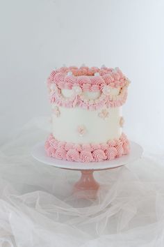 a white and pink cake with ruffles on top sitting on a plate in front of a white backdrop