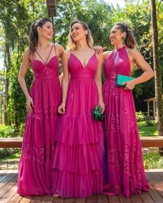 three women in pink dresses standing together on a wooden deck with trees in the background