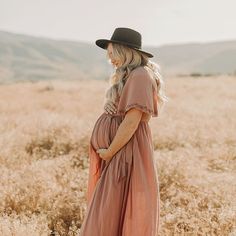 a pregnant woman standing in a field wearing a black hat and pink dress with long sleeves