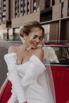 a woman standing next to a red car wearing a white wedding dress and holding a veil