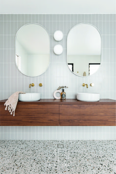 two round mirrors on the wall above sinks in a white tiled bathroom with wood cabinets