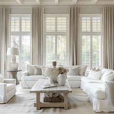 a living room filled with white furniture and windows covered in shades of beige drapes