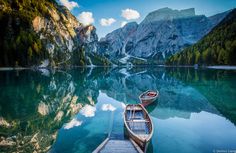 two boats are docked on the water in front of some mountains and pine trees with clouds