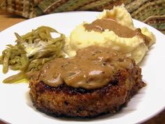 a white plate topped with meat covered in gravy next to mashed potatoes and green beans