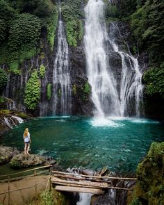 a woman standing in front of a waterfall with blue water and green trees surrounding it