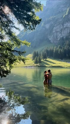 two people are sitting on the edge of a body of water with mountains in the background