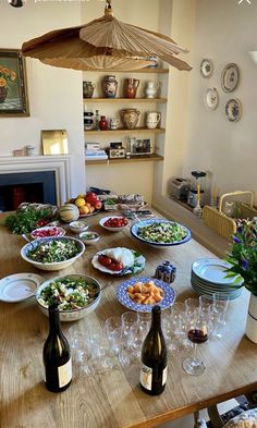 a wooden table topped with lots of plates and bowls filled with food next to bottles of wine