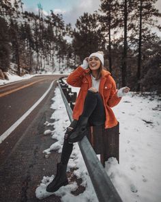 a woman in an orange jacket sitting on a fence next to snow covered ground and trees