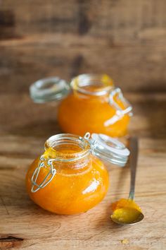 two jars filled with orange marmalade sitting on top of a wooden table next to a spoon