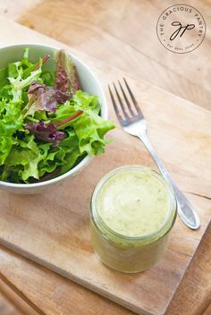 a salad in a bowl next to a jar of dressing on a cutting board with a knife and fork