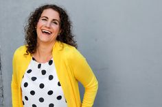 a woman with curly hair wearing a yellow cardigan and polka dot top smiles at the camera